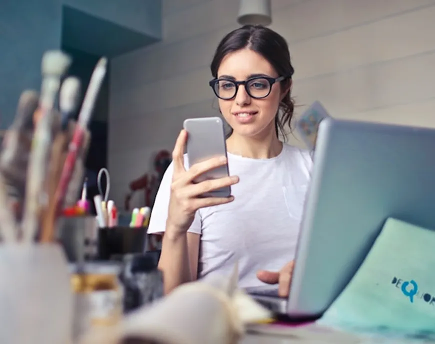 A woman wearing glasses and a white T-shirt, holding a smartphone while sitting at a desk with an open laptop. Art supplies, including brushes and pens, are visible in the foreground, suggesting a creative workspace.