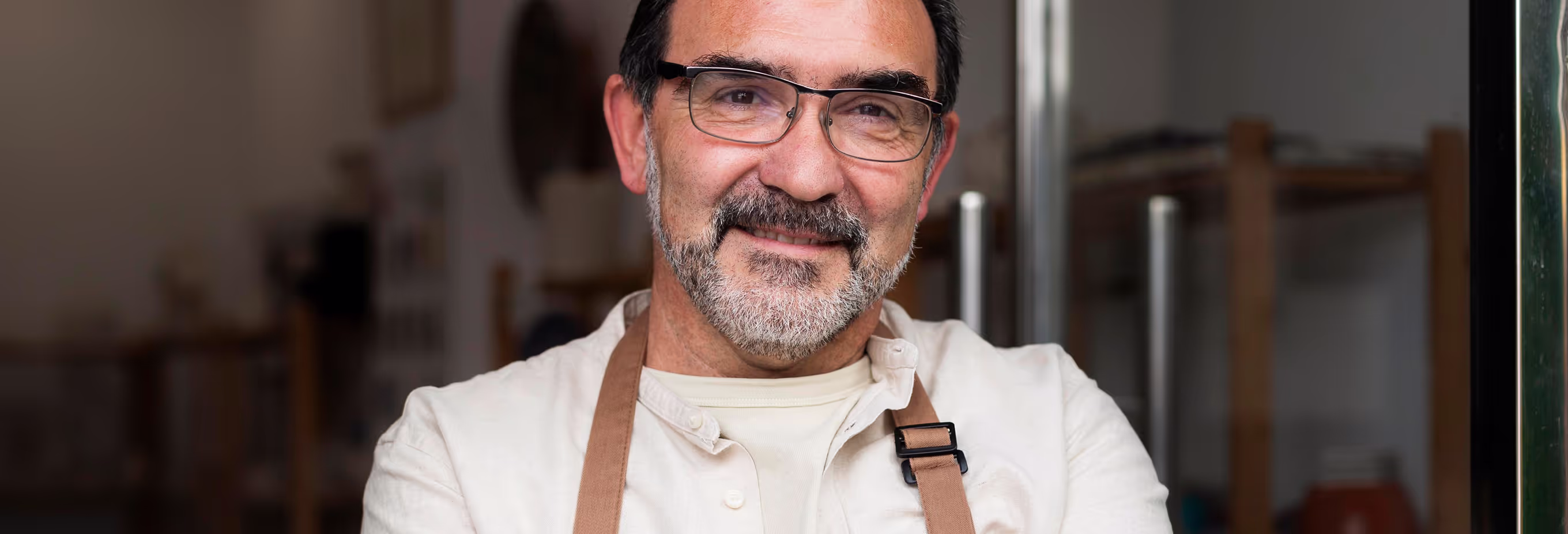 A close-up portrait of a smiling man with glasses and a neatly trimmed beard, wearing a beige shirt and a brown apron. The background suggests a cozy, indoor workspace or studio.