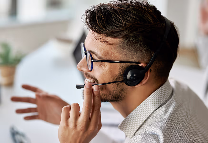 A person in profile wearing a headset and glasses, gesturing with one hand while smiling during what appears to be a professional call. They have short dark hair and are wearing a light patterned shirt in a bright office environment.