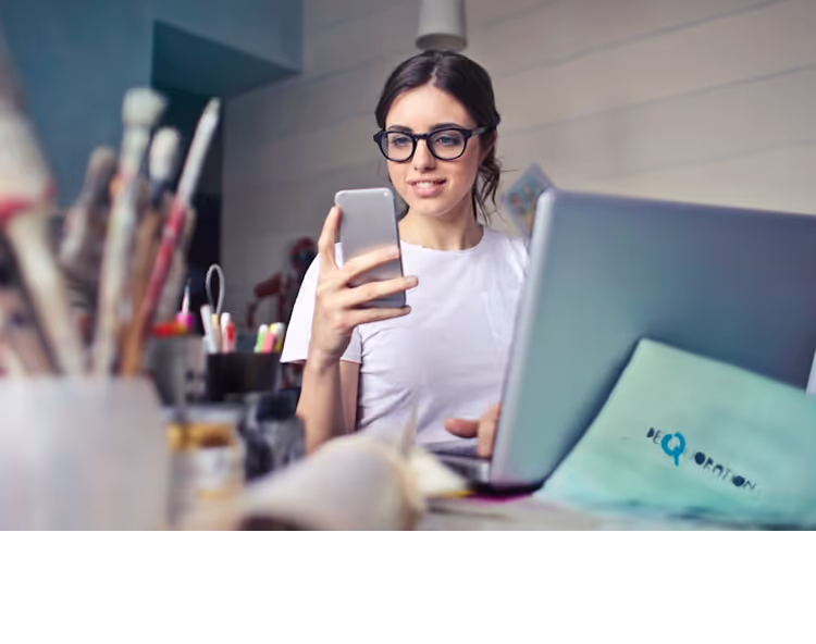 A woman wearing glasses and a white T-shirt, holding a smartphone while sitting at a desk with an open laptop. Art supplies, including brushes and pens, are visible in the foreground, suggesting a creative workspace.