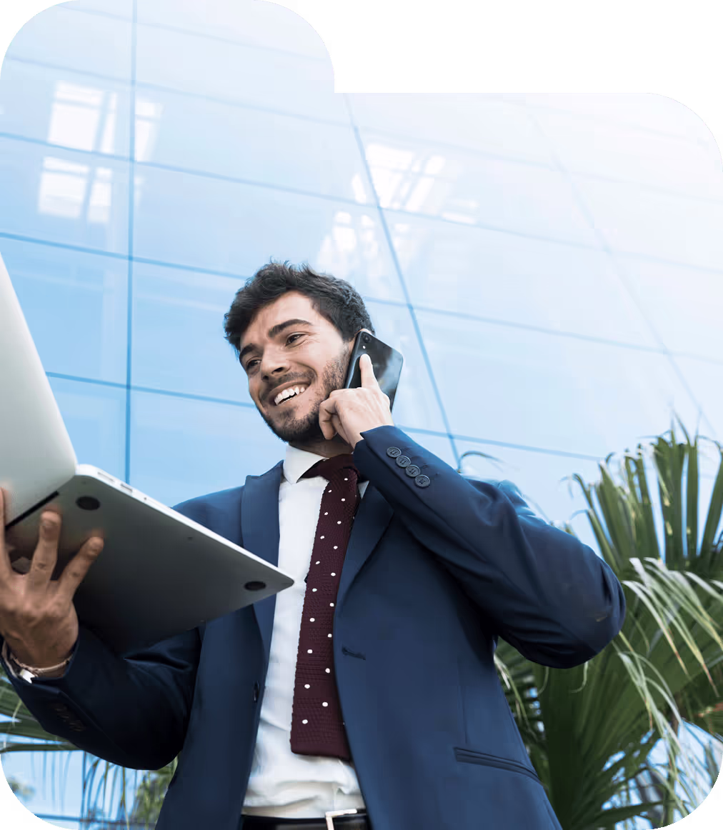 The image shows a smiling businessman in a navy blue suit with a white shirt and burgundy tie with white dots. He is holding a laptop in one hand while talking on a smartphone with the other. He appears to be outside a modern building with glass facades, and there are some green plants visible in the background. The low angle perspective and glass building suggest a professional, corporate environment. The man looks happy and engaged in his conversation while multitasking with his devices.