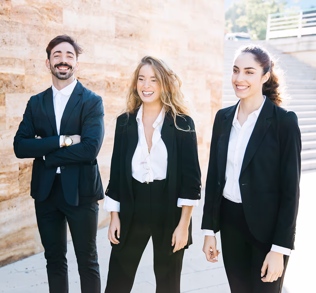 Three professionals wearing formal black suits and white shirts, standing outdoors and smiling confidently. The group consists of two women and one man, all exuding a positive and approachable demeanor.